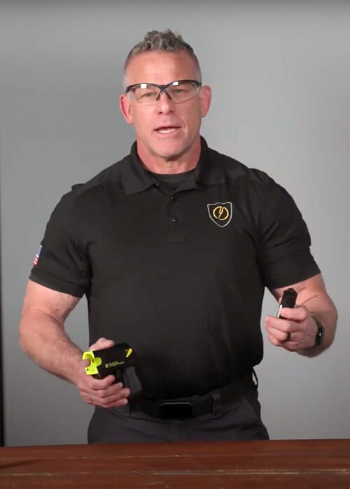 A man standing behind a table wearing safety glasses and is holding a TASER device
